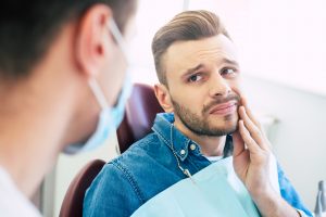 Symptoms Of Gums Pain. A Man With A Worried Face Is Holding His Hand On His Cheek Because Of Irritating Pain In Front Of A Dentist Who Is Going To Give A Patient A Treatment.