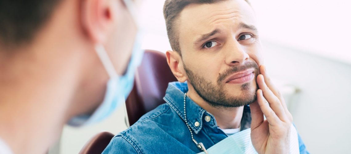 Symptoms Of Gums Pain. A Man With A Worried Face Is Holding His Hand On His Cheek Because Of Irritating Pain In Front Of A Dentist Who Is Going To Give A Patient A Treatment.